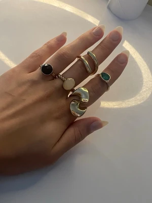 Mixade guldringar med stenar - Säljer ett set med fem trendiga guldfärgade ringar i olika former. Några ringar har färgade stenar i svart, vitt och grönt, medan andra har en chunky, vågig design. Perfekta för att skapa en personlig och unik stil.