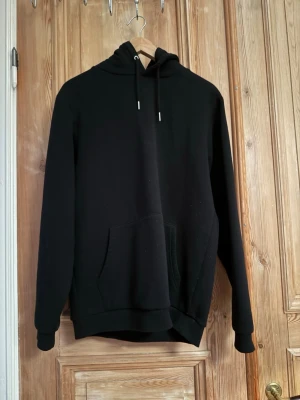 Svart hoodie med ficka och huva - En klassisk svart hoodie med stor magficka och justerbar huva med snören. Hoodien har ribbade muddar vid ärmar och nederkant, perfekt för en avslappnad och stilren look. Passar till det mesta och är superenkel att matcha med andra plagg.