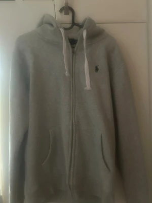 Grå zip hoodie från Polo Ralph Lauren - Snygg grå hoodie med dragkedja från Polo Ralph Lauren. Klassisk design med huva och vita snören, två fickor framtill och den ikoniska loggan broderad på bröstet. Perfekt till chill dagar och enkel att matcha med allt.