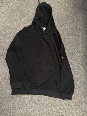 Svart hoodie från Stone Island - Säljer en svart hoodie från Stone Island med klassisk patch på vänster ärm. Hoodien har huva med dragsko, magficka och ribbade muddar. Perfekt för dig som gillar streetwear och vill ha en stilren look.