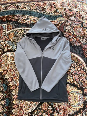 Grå hoodie från Under Armour - Snygg tvåfärgad hoodie från Under Armour i ljusgrått och mörkgrått. Dragkedja framtill, huva med logga och två sidofickor. Tillverkad i ett mjukt och stretchigt material som passar perfekt för en  look. För 579kr