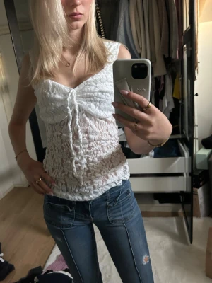 Vit spetslinne med snörning - Säljer ett vitt, ärmlöst spetslinne med snygg snörning framtill. Toppen är genombruten och har en v-ringad form som ger en feminin look. Perfekt att styla med jeans eller kjol för en trendig outfit.