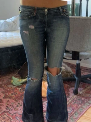 Mörkblå slitna bootcut jeans - Säljer ett par mörkblå bootcut jeans med slitningar och stora hål vid knäna. Jeansen har låg midja, klassiska fem fickor och en tvättad look. Perfekta för dig som gillar en edgy och avslappnad stil.