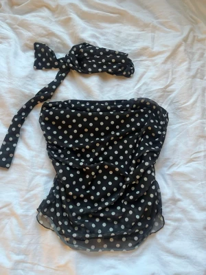 Svart bandeau top med vita prickar. - Säljer en svart bandeau tubtopp med vita prickar och har en prickig scarf till. Toppen har rynkade detaljer framtill och ärmlös design. Materialet känns som ett lätt och stretchigt syntetmaterial, perfekt för en retroinspirerad look.