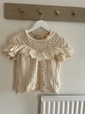 Beige stickad topp med volang - Supersöt beige stickad topp från Zara med korta ärmar och bred volangdetalj över bröstet och axlarna. Toppen har ett genombrutet mönster och ribbad halsringning. Perfekt för dig som gillar romantisk och trendig stil. Är storlek M men passar mer som en xs. 