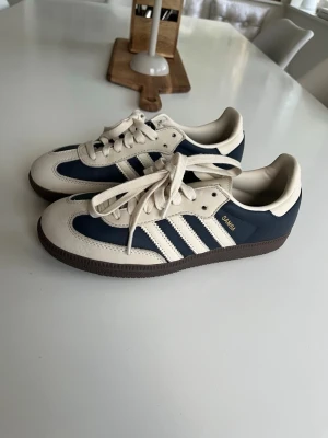 Adidas Samba OG blåvita sneakers Bra skick! - Snygga Adidas Samba OG sneakers i blått och vitt med klassiska tre ränder på sidan och gummisula. Ovandelen är i läder och mocka, med gulddetalj på sidan. Perfekta för dig som gillar retrostil och streetwear.
