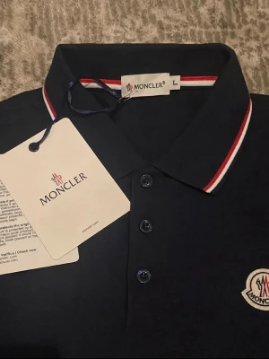 Mörkblå Moncler pikétröja storlek L/3 - Snygg mörkblå pikétröja från Moncler med klassisk krage och knäppning med tre knappar. Kragen har vita och röda ränder och bröstet pryds av Monclers ikoniska logotyp. Perfekt för en stilren och sportig look.