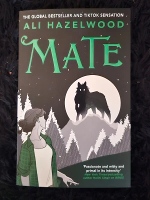 Mate av Ali Hazelwood - En engelskspråkig skönlitterär bok med svart och grön framsida. Omslaget visar en tecknad kvinna i grön skjorta och en varg mot en månbelyst skog. Boken är en global bestseller och populär på TikTok.