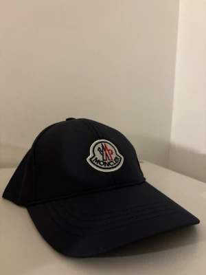 Svart keps från Moncler - Snygg svart keps från Moncler med klassisk böjd skärm och broderad Moncler-logga framtill i vitt, rött och blått. Kepsen har en stilren design och är tillverkad i ett slätt material som ger en modern look. 