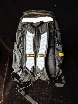 Svart Nike Elite ryggsäck - Snygg svart Nike Elite ryggsäck med gula detaljer och logga. Ryggsäcken har vadderade axelband med reflekterande partier, flera fack med dragkedja och sidofickor. Tillverkad i slitstarkt material, perfekt för skola eller träning.