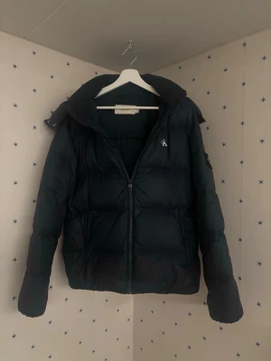 Mörkgrön pufferjacka med huva - Säljer en mörkgrön pufferjacka med huva och dragkedja framtill. Jackan har en broderad logga på bröstet och två sidofickor. Puffig design som håller värmen under kalla dagar. Perfekt för dig som vill ha en stilren och funktionell vinterjacka.