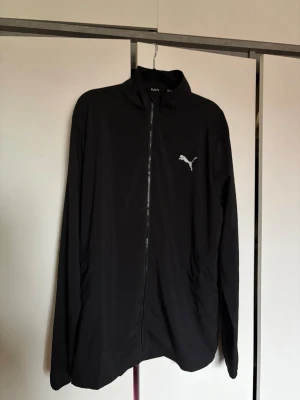Svart vindjacka från Puma - Snygg svart vindjacka från Puma med hel dragkedja framtill och diskret Puma-logga på bröstet. Jackan är tillverkad i lätt, vindtätt material som passar perfekt för löpning eller andra utomhusaktiviteter.