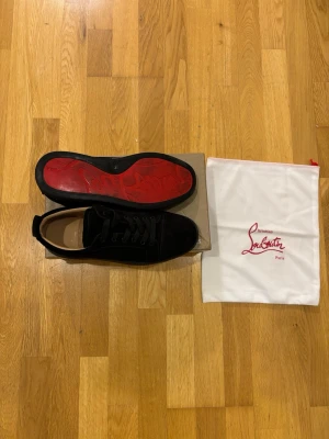 Svarta loubs  - Stilrena svarta sneakers från Christian Louboutin i mocka med klassisk röd sula. Skorna har lågt skaft, svarta skosnören och diskret design som passar till många olika outfits. Medföljer original dustbag.