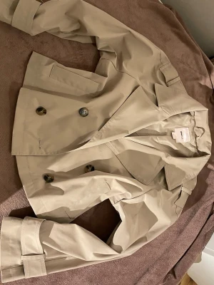 Kort beige trenchcoat med dubbelknäppning - Stilren kort trenchcoat i beige med dubbelknäppning och bred krage. Jackan har axelklaffar, sidofickor och justerbara remmar vid ärmsluten. Tillverkad i ett slätt tyg som ger en klassisk look. Perfekt för dig som gillar tidlös och trendig stil.