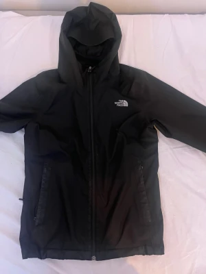 Svart vindjacka från The North Face - Snygg svart vindjacka från The North Face med huva och dragkedja framtill. Jackan har två fickor med dragkedja, elastiska muddar vid ärmsluten och logga på bröstet. Tillverkad i lätt och vattenavvisande material, perfekt för blåsiga dagar.                                                                     (Använd)