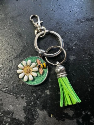 Nyckelring med blomma och bi - Snygg nyckelring med en stor vit blomma och ett litet bi på en grön bakgrund. Nyckelringen har även en grön tofs i mockaimitation och metallfäste. Perfekt att hänga på väskan eller nycklarna för en färgglad touch.