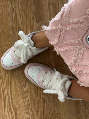 Nike sneakers i vitt och rosa - Säljer ett par snygga Nike sneakers i vitt och ljusrosa med fluffiga, breda snören. Skorna har klassisk perforerad tå och lågt skaft. Perfekta för dig som vill ha en unik twist på dina sneakers.