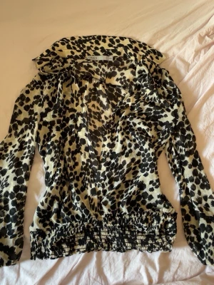 Leopardmönstrad blus från Oasis - Snygg blus från Oasis med svartvitt leopardmönster. Blusen har lång ärm, bred krage och resår i midjan för en skön passform. Tillverkad i ett lätt, glansigt material som ger en cool look.