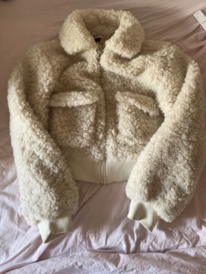 Krämvit teddyjacka med fickor - Mysig krämvit teddyjacka med fluffigt material och två stora bröstfickor. Jackan har dragkedja framtill, bred krage och ribbade muddar vid ärmslut och nederkant. Perfekt för kalla dagar när du vill vara både varm och trendig.