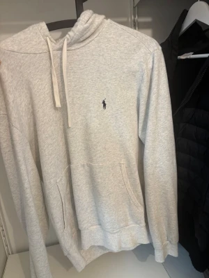 Ljusgrå hoodie från Polo Ralph Lauren - Ljusgrå hoodie med klassisk huva och dragsko från Polo Ralph Lauren. Har en broderad marinblå logga på bröstet och en stor känguruficka framtill. Tillverkad i mjukt sweatshirtmaterial och har ribbade muddar vid ärmslut och nederkant.