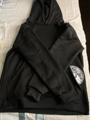 Svart hoodie med patchdetalj - Säljer en svart hoodie med stor huva och ribbade muddar. På ena ärmen finns en tydlig patch med text och grafisk design. Hoodien har en klassisk känguruficka framtill och är gjord i mjukt material, perfekt för en avslappnad stil.