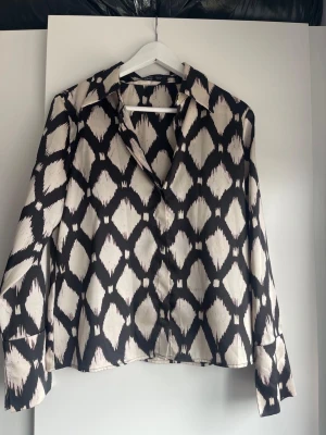 Svartvit mönstrad skjorta i satinlook - Snygg skjorta med svart och vit diamantmönster och glansig satinliknande yta. Skjortan har lång ärm, klassisk krage och knäppning framtill. Perfekt för dig som vill sticka ut med ett coolt mönster och skön passform.