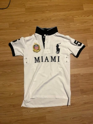 Vit Polo Ralph Lauren Miami t-shirt - Vit t-shirt från Polo Ralph Lauren med svart krage och detaljer. Stor MIAMI-text framtill, broderad patch och klassiska polospelaren-loggan. Kortärmad modell med knappar vid halsen och kontrastfärgade muddar. Perfekt för en sportig och stilren look.