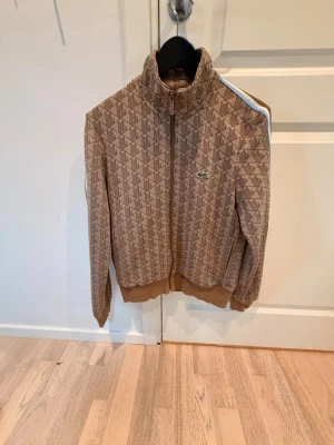 Beige mönstrad jacka från Lacoste - Säljer en beige jacka från Lacoste med brunt geometriskt mönster och dragkedja framtill. Jackan har hög krage, ribbade muddar och den klassiska gröna krokodilloggan på bröstet. Perfekt för dig som gillar sportig och retro stil.