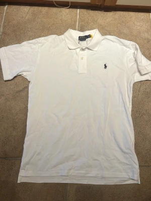 Vit Piké från Polo Ralph Lauren - Klassisk vit pikétröja från Polo Ralph Lauren med den ikoniska broderade loggan på bröstet. Tröjan har krage, korta ärmar och knäppning med två knappar framtill. Perfekt till jeans eller shorts för en clean och stilren look. Storleken sitter lite mindre, som L. Skriv vid frågor:)