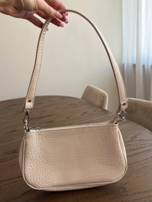 Beige axelremsväska - Stilren beige handväska med axelrem och krokopräglat mönster. Väskan har en avlång form, silverfärgade metalldetaljer och dragkedja upptill. 