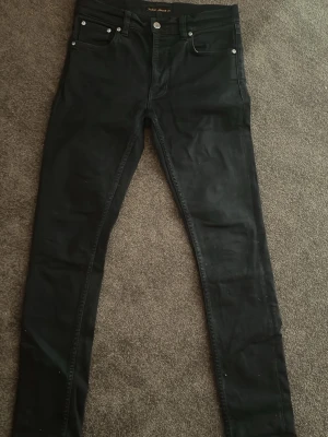 Svarta jeans från Nudie Jeans - Säljer ett par svarta jeans från Nudie Jeans. Klassisk femficksmodell med silverfärgade nitar och knapp. Jeansen har smal passform och är tillverkade i ett stretchigt denimtyg för extra komfort.