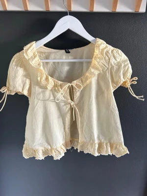 Söt ljusgul blus med spets och knyt - Supersöt ljusgul blus med volangkant, spetsdetaljer och knyt fram. Puffiga korta ärmar med knytband och romantisk känsla. Perfekt till jeans eller kjol för en gullig look.