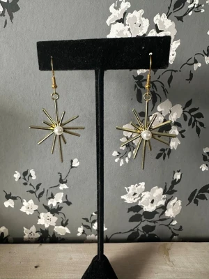 Guldiga stjärnörhängen med pärldetalj - Unika örhängen i form av stjärnor i guldfärgad metall med en vit pärla i mitten. Hängande modell med krokfäste som ger en cool och trendig look. Perfekta för dig som vill sticka ut med accessoarer som har det lilla extra.