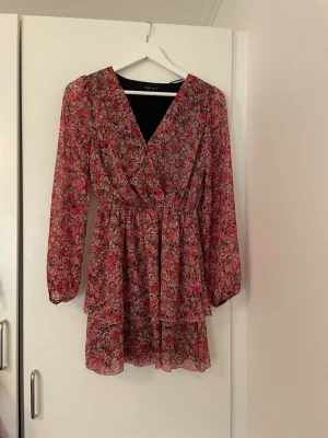 Blommig kortklänning med v-ringning - Söt kortklänning med blommigt mönster i rosa, rött och vitt. Klänningen har v-ringning, långa ärmar och markerad midja med lätt volang i kjolen.