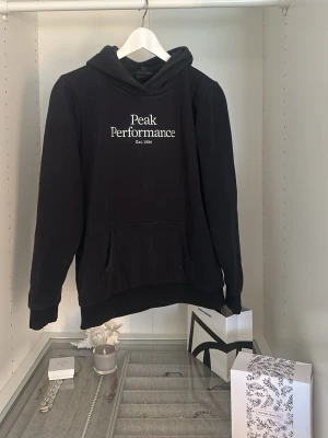 Svart hoodie Peak Performance - Säljer en svart hoodie från Peak Performance med vit broderad logga på bröstet. Klassisk känguruficka framtill och mjuk insida. Perfekt för dig som gillar en enkel och stilren look. Dragsko i huvan och ribbade muddar vid ärmar och nederkant.