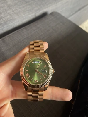 Klocka - Stilren klocka i guldtonat metallarmband och boett. Urtavlan är grön med romerska siffror och dag- samt datumvisning. Klockan har en klassisk länkdesign och markerad räfflad kant runt urtavlan. Perfekt accessoar för dig som gillar detaljer.
