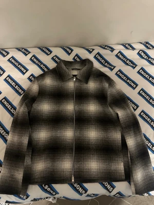 Gråmelerad overshirt från H&M - Snygg gråmelerad overshirt från H&M med diskret rutmönster. Jackan har dragkedja framtill, klassisk krage och raka ärmar. Perfekt lager på lager-plagg för en stilren look. Materialet känns mjukt och har en ull-liknande känsla.