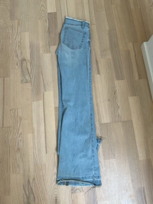 Ljusblå bootcut jeans med rå kant - Säljer ett par ljusblå bootcut jeans med rå, fransig kant nedtill. Jeansen har klassisk femficksdesign och är tillverkade i denim med en avslappnad passform. Perfekta för en casual och trendig look.