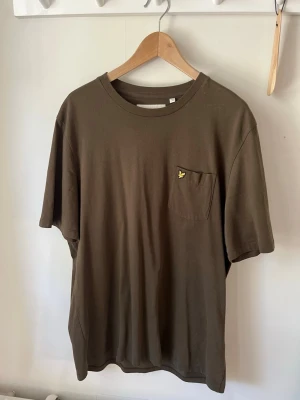 Brun t-shirt med bröstficka - Säljer en enkel brun t-shirt med rund hals och bröstficka. På fickan sitter en liten gul broderad logga. T-shirten har normal passform och är tillverkad i mjuk bomull, perfekt för en avslappnad stil.