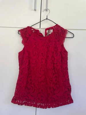 Röd spetsblus med ärmlös design - Snygg röd blus i spets med ärmlös design och rund hals. Blusen har fina spetsdetaljer över hela plagget och en feminin passform. 