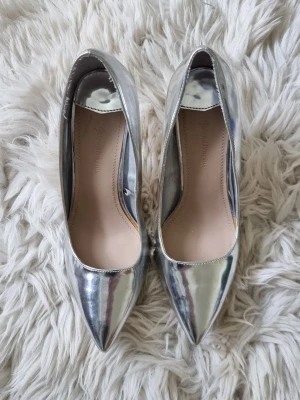 Silvriga spetsiga pumps med klack - Säljer ett par glansiga silvriga pumps med spetsig tå och hög smal klack. Skorna har en stilren design och är tillverkade i ett syntetiskt material med metallic-finish. Perfekta för dig som vill sticka ut med en futuristisk look. En repa finns på ena sidan (se bild)