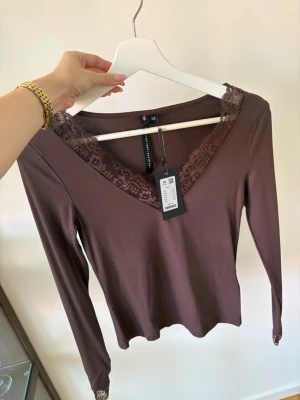 Brun långärmad topp med spets - Snygg brun långärmad topp med v-ringning och spetskant runt halsen och handlederna. Tillverkad i mjukt material som känns skönt mot huden. 