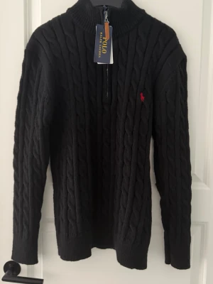 Svart kabelstickad tröja Polo Ralph Lauren - Kabelstickad svart tröja från Polo Ralph Lauren med halv dragkedja och hög krage. Tröjan har ett rött broderat logotyp på bröstet och ribbstickade muddar. Perfekt för dig som gillar klassisk och stilren design.