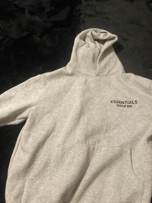 Grå Essentials Fear of God hoodie Storlek S - Snygg grå hoodie från Essentials Fear of God med svart logga tryckt på bröstet och ryggen. Klassisk känguruficka framtill och stor huva. Tillverkad i mjukt sweatshirtmaterial som är perfekt för en avslappnad streetwear-look.