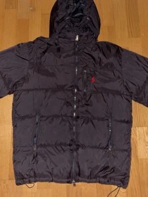 Svart jacka från ralph Lauren - Snygg svart dunjacka med huva och dragkedja framtill från Ralph Lauren. Jackan har två sidofickor med dragkedja och en bröstficka med dragkedja. På bröstet finns en liten röd Ralph Lauren logga. Perfekt för kalla vinterdagar. Liten i storlek.