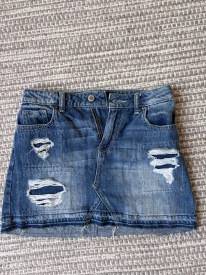 Blå jeanskjol med slitningar - Säljer en blå jeanskjol i kort modell med slitna detaljer och rå kant nertill. Kjolen har klassiska fickor fram och bak samt bälteshällor. Perfekt för dig som gillar en avslappnad och trendig stil.