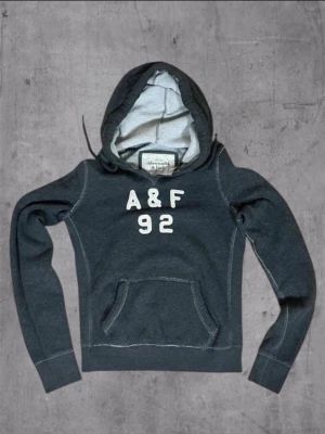 Mörkgrå hoodie Abercrombie & Fitch - Mörkgrå hoodie med huva och magficka från Abercrombie & Fitch. Framtill står det 'A&F 92' i vita bokstäver. Hoodien har ribbade muddar och kontrastsömmar samt mjukt innerfoder. Perfekt för en avslappnad och sportig stil. Kan diskutera pris🤝