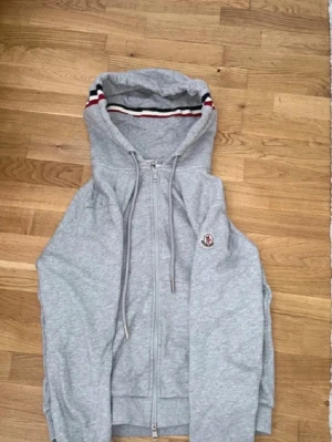 Grå Moncler hoodie med dragkedja - Snygg grå hoodie från Moncler med dragkedja framtill och huva med röd, vit och marinblå rand på insidan. Klassisk Moncler-logga på bröstet. Tillverkad i mjukt bomullsmaterial och har dragsko i huvan för extra stilpoäng.