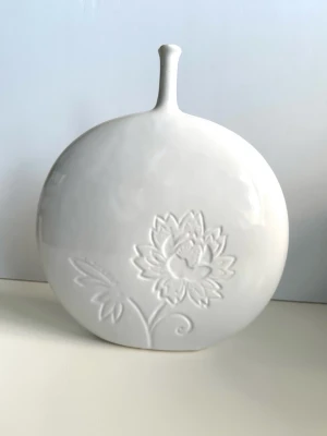 Vit dekorativ keramikvas med blomrelief - Stilig vit dekorativ vas har ett elegant och diskret blommotiv både fram och bak. Vacker modell med stor rund front och smal pip i toppen. Observera att den har en liten spricka på ena sidan, se bild, men ingenting som man tänker på när den står framme (särskilt inte om man vänder den med andra sidan fram). 25 cm bred och 27 cm hög. Mångsidig inredningsdetalj som passar till alla rum. Lika fin som dekoration precis som den är eller med torkade blommor i. Fin!