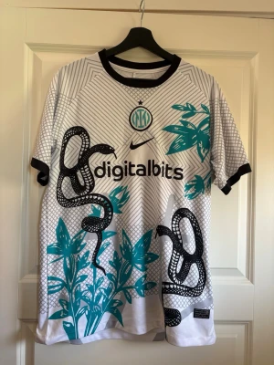 Inter Milan fotbollströja  - Säljer en vit Inter Milan matchtröja från Nike med svarta och turkosa detaljer. Tröjan har ett unikt ormmotiv och växtgrafik, svarta ränder och klubbmärke på bröstet. Tillverkad i lätt och ventilerande material, perfekt för fotboll eller träning.